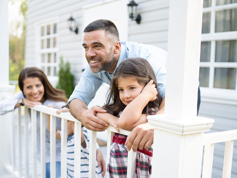 a father, mother, and daughter stand on a white porch attached to their white house