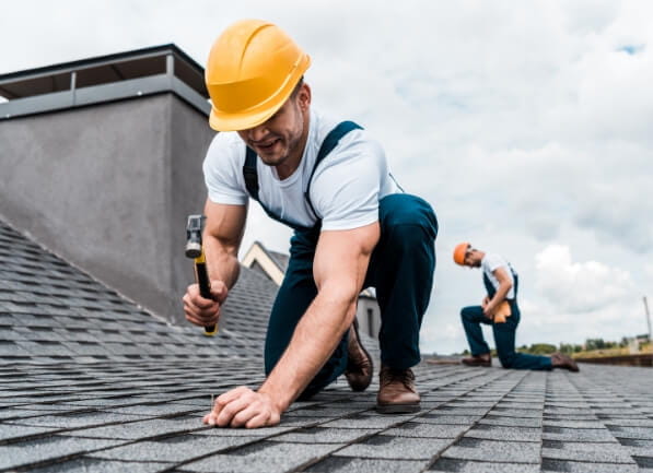 two roofing contractors with yellow hard hats and hammering shingles into the roof of a house